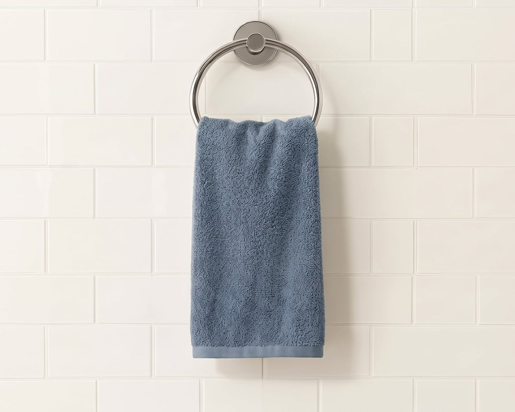 A blue-gray cotton hand towel hangs from a round silver metal towel ring mounted on a white subway tile wall.