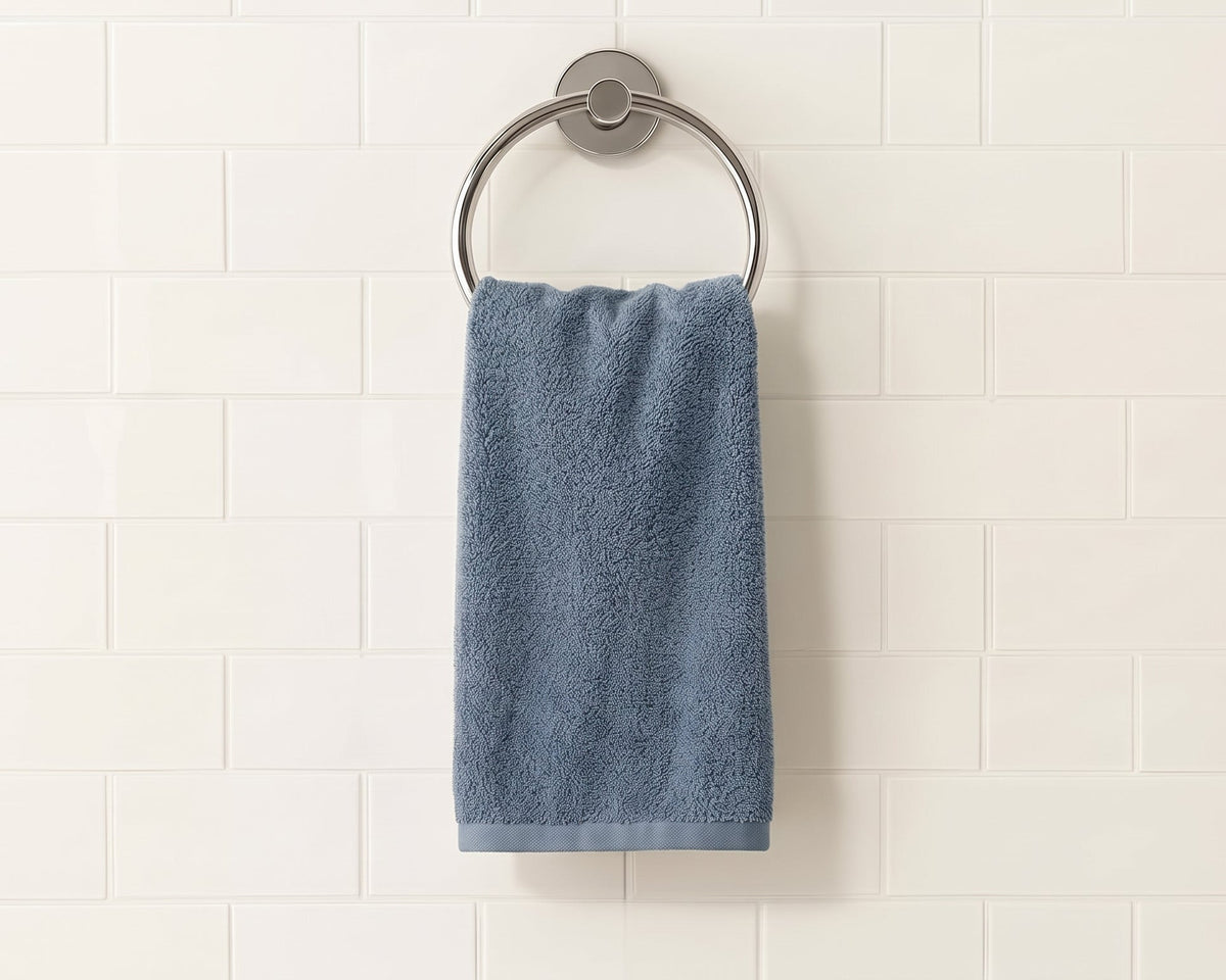A blue-gray cotton hand towel hangs from a round silver metal towel ring mounted on a white subway tile wall.