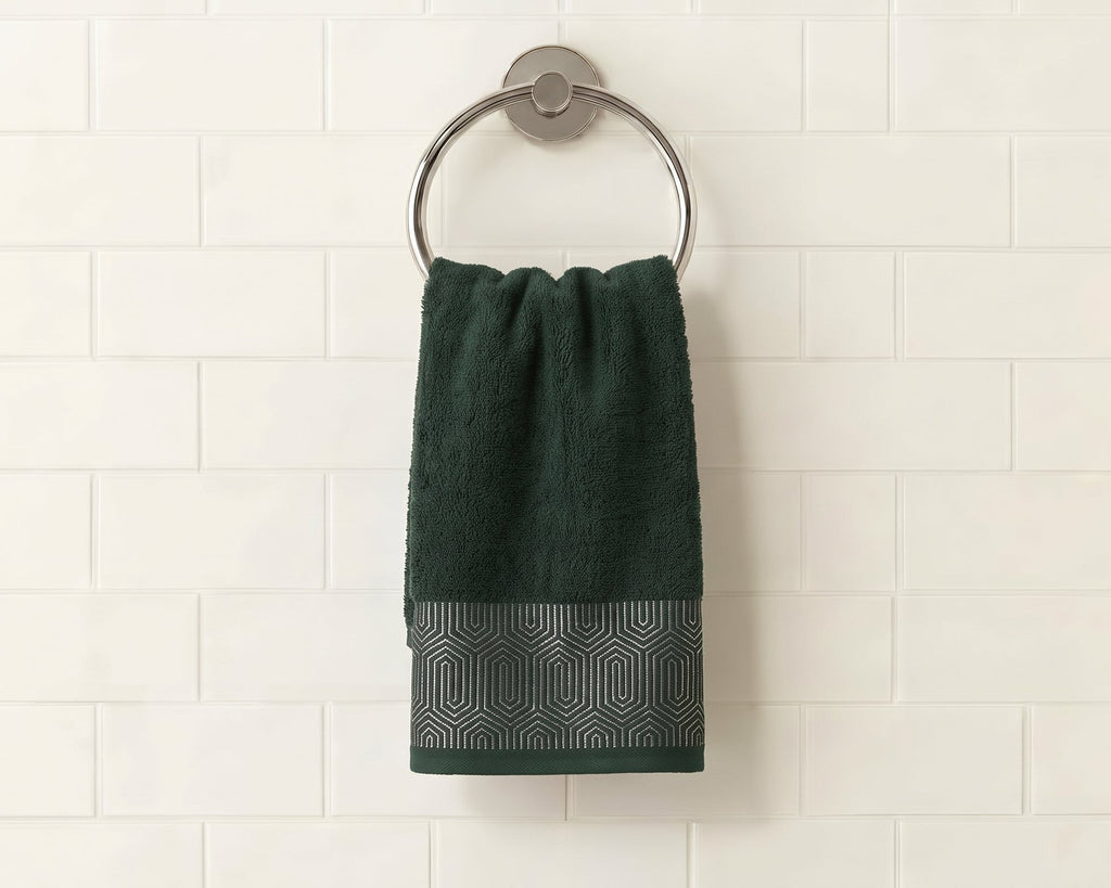 A dark green hand towel with a textured geometric pattern border hangs from a round silver metal towel ring on a white subway tile wall.