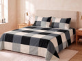 A neatly made bed in a softly lit bedroom, featuring a large-scale buffalo plaid bedding set in black, white, and gray. The set includes a flat sheet, a comforter/duvet, and two matching pillowcases.