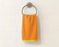 An orange hand towel with a lime green patterned border hangs from a round silver metal towel ring mounted on a white subway tile wall.